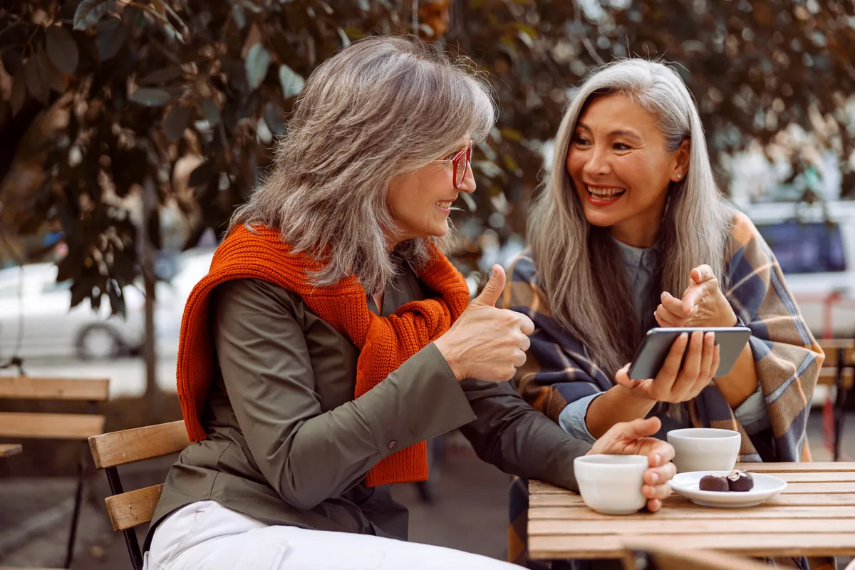Two women with gray hair laughing together at an outdoor cafe table, one giving a thumbs up while the other holds a phone