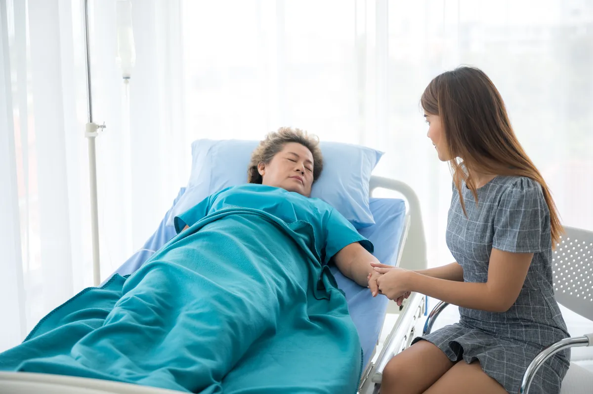 Younger woman in a gray dress sitting beside a hospital bed and holding the hand of an older woman who is lying under a teal blanket with her eyes closed, in a bright room with white curtains