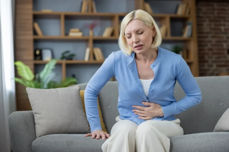 Woman in her 50s sitting on a sofa with a hand on her abdomen, looking uncomfortable