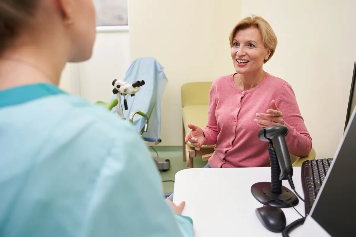 Smiling woman with short blonde hair and a pink cardigan gesturing as she talks across a desk with a clinician in a light blue top