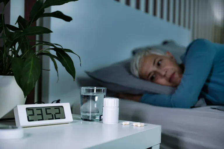 Woman with gray hair lying awake in bed at night, looking toward a bedside table with a digital clock showing 23:52, a glass of water, a pill bottle and two capsules