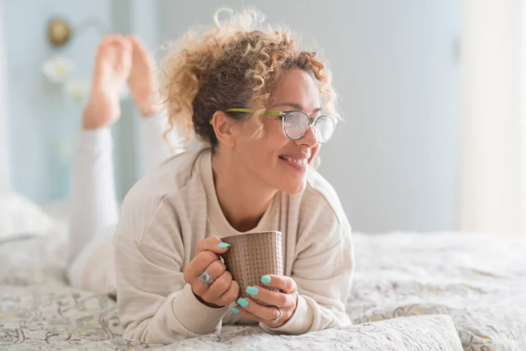 Woman with curly hair and glasses relaxing in bed with a mug