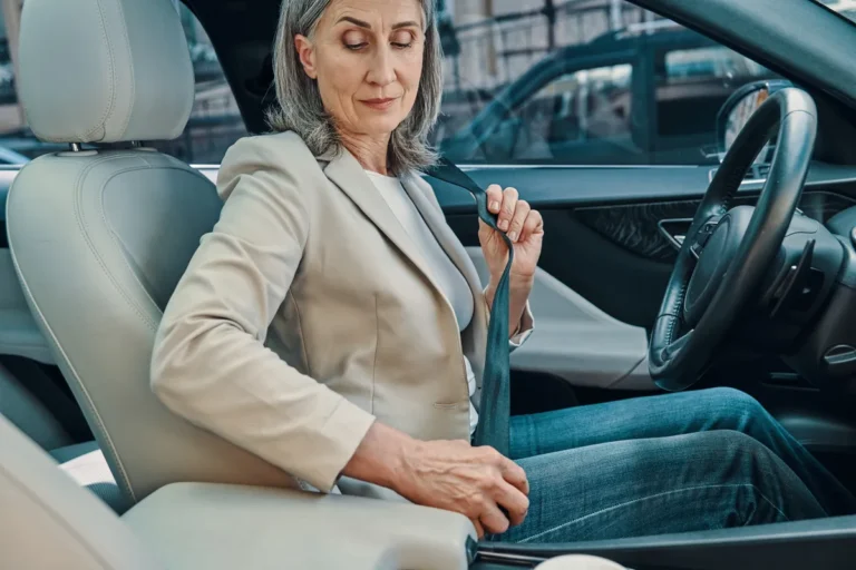 Woman with shoulder-length grey hair in a beige blazer fastening her seatbelt in the front seat of a car