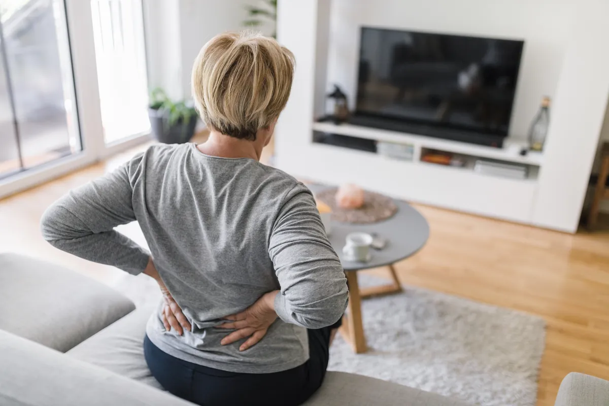 Woman seen from behind, sitting on a couch with both hands pressed to her lower back
