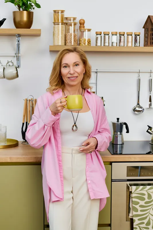 Smiling woman with shoulder-length blond hair in a pink shirt holding a yellow-green mug in a kitchen