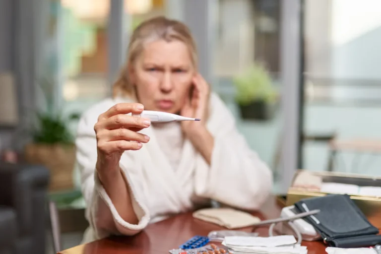 Woman in a white bathrobe holding up a digital thermometer at a table with a blood pressure monitor and medicines
