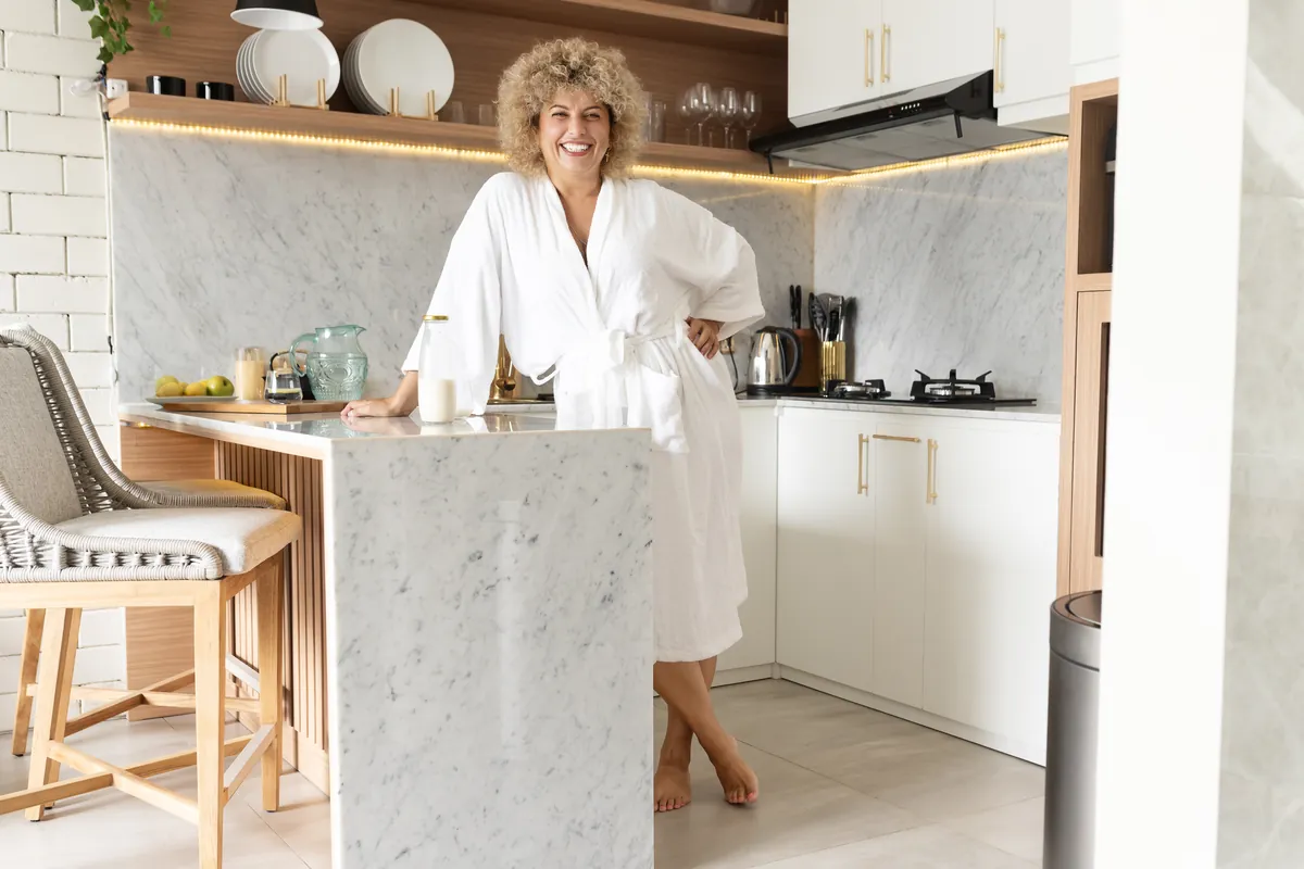 Smiling woman with curly blond hair in a white bathrobe leaning on a marble kitchen counter at home