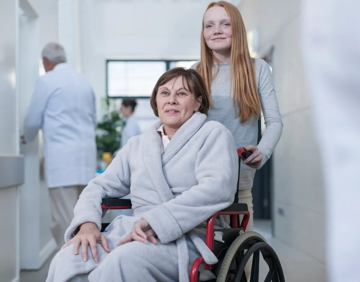 Woman in a light grey fleece robe sitting in a wheelchair in a hospital corridor, pushed by a smiling red-haired teenager