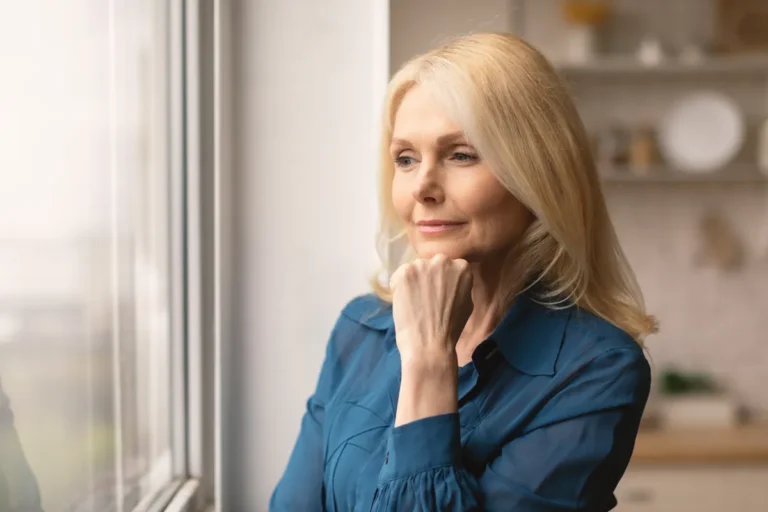 Woman with shoulder-length blond hair and a blue blouse looking thoughtfully out of a window with her hand under her chin