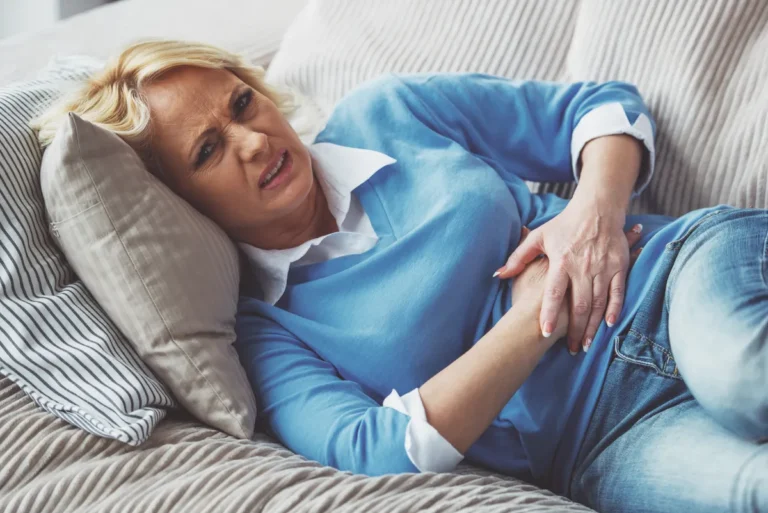Middle-aged blonde woman in a blue sweater lying on a couch with a pained expression, holding her lower abdomen