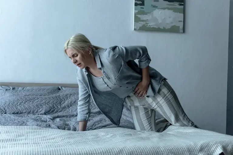 Woman in pajamas standing by her bed with one hand on her lower abdomen