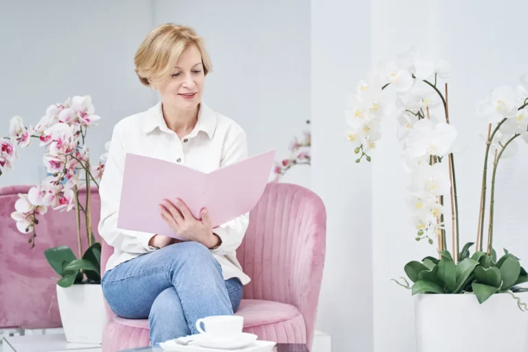 Woman in her fifties with short blond hair, a white shirt and jeans, sitting in a pink armchair reading a pink folder, with white and pink orchids around her