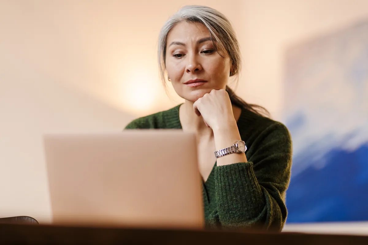 Woman with long grey hair and a green sweater resting her chin on her hand while reading something on a laptop