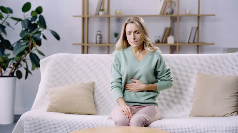 Blonde woman in a mint sweater sitting on a sofa with a hand on her abdomen, looking uncomfortable