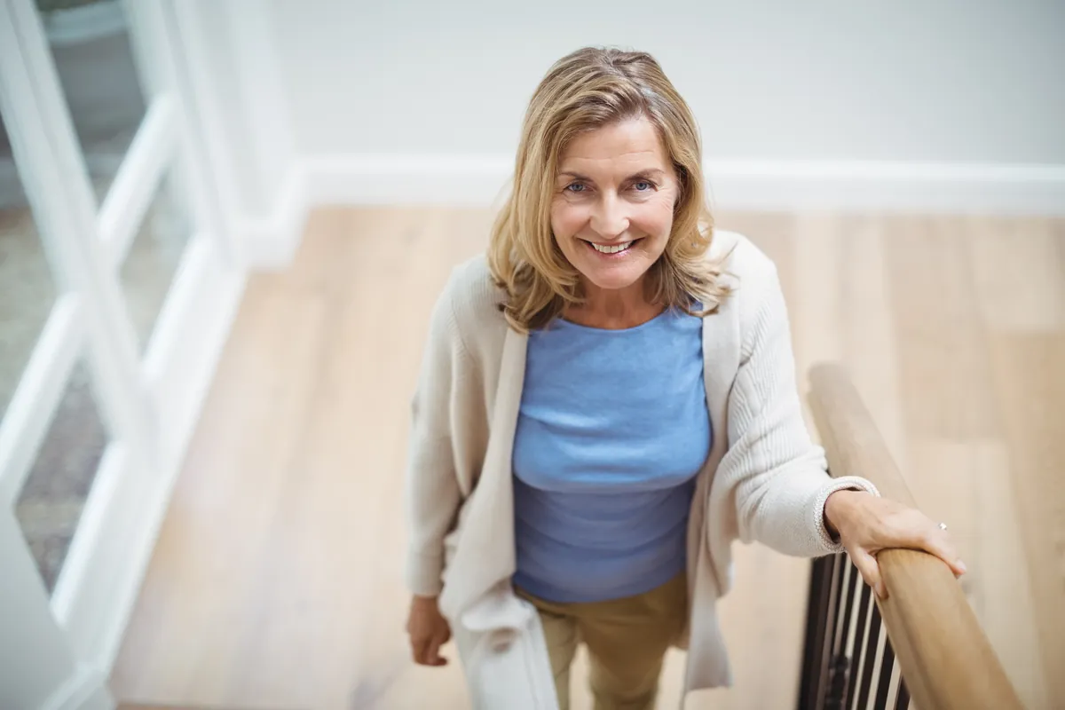 Smiling woman with shoulder-length blond hair in a cream cardigan walking up a wooden staircase with her hand on the banister
