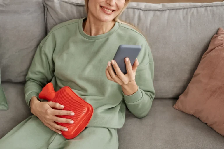 Woman in a sage green sweatsuit sitting on a grey sofa holding a red hot water bottle against her lower abdomen and a phone in her other hand
