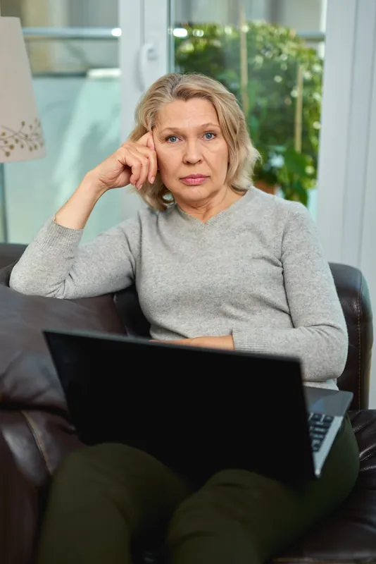 Woman with wavy blonde hair and a grey sweater sitting on a leather sofa with a laptop on her knees, resting her head on her hand