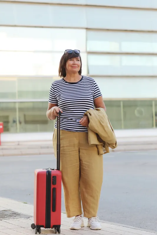Woman in her fifties in a striped T-shirt and beige trousers standing on a sidewalk outside a glass-fronted building with a small red carry-on suitcase