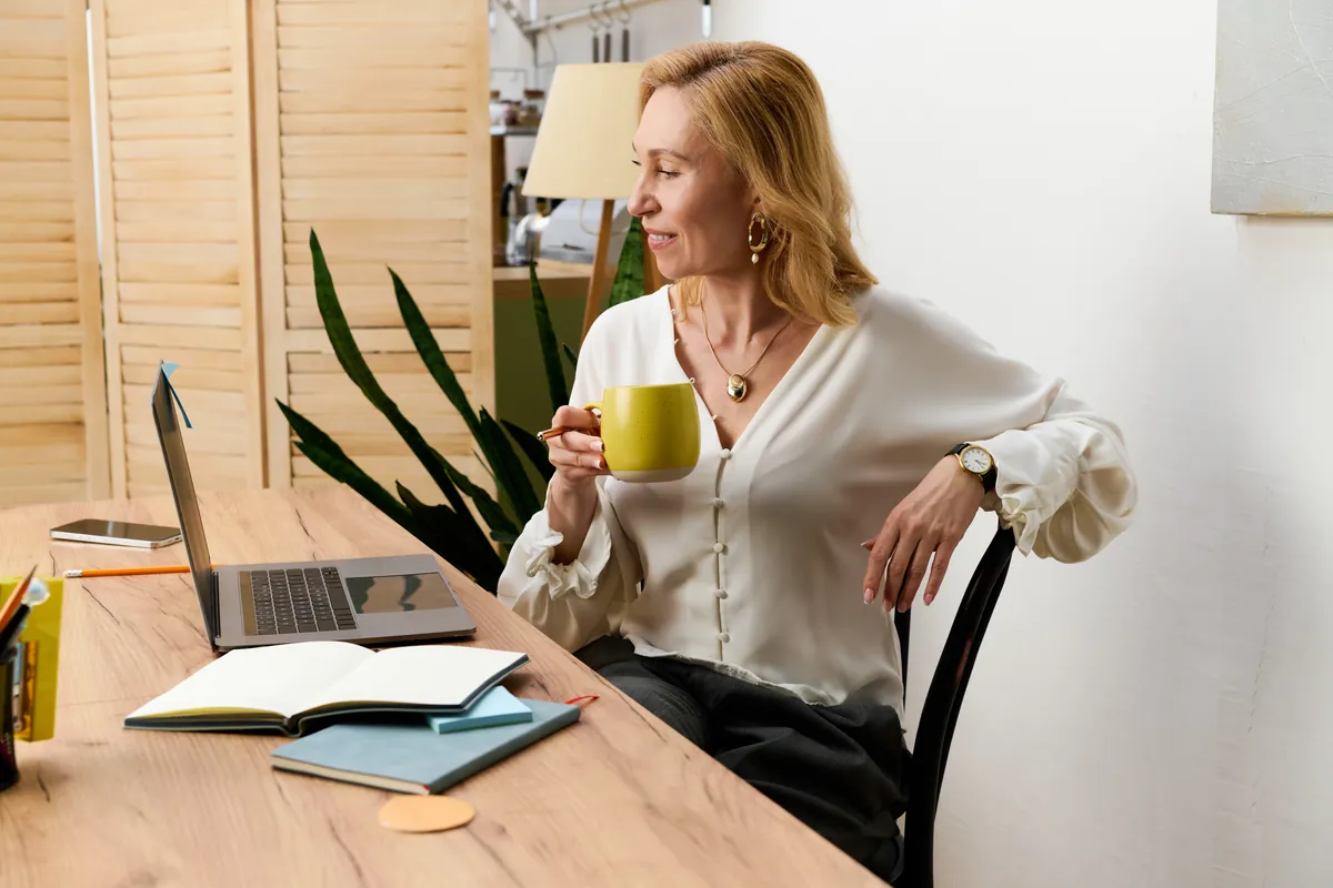 Woman with shoulder-length blond hair in a white blouse holding a yellow mug, sitting at a wooden desk with an open laptop, notebooks and a phone