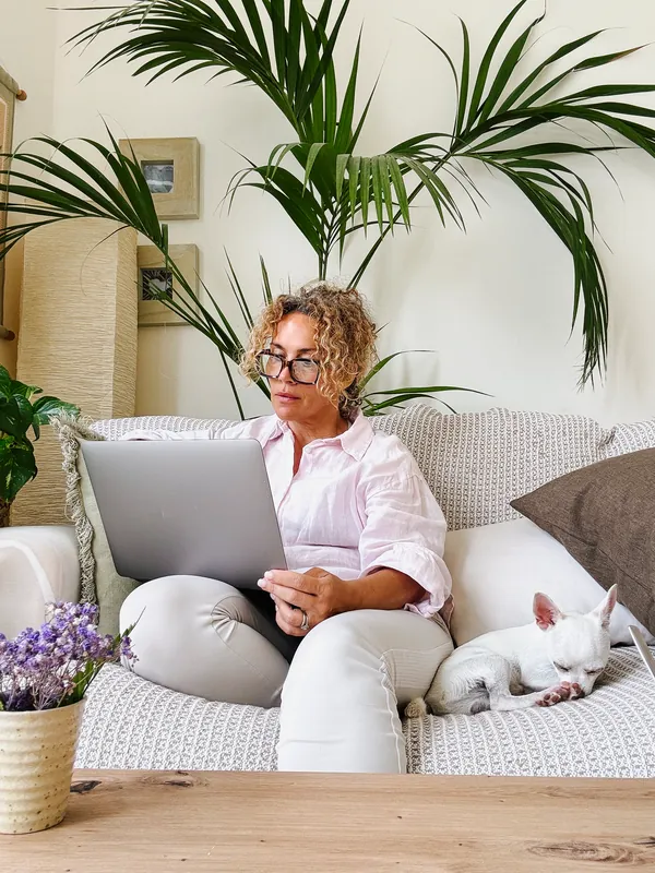 Woman with curly hair and glasses in a light pink shirt working on a laptop on a sofa, with a small white dog asleep next to her and a palm plant behind