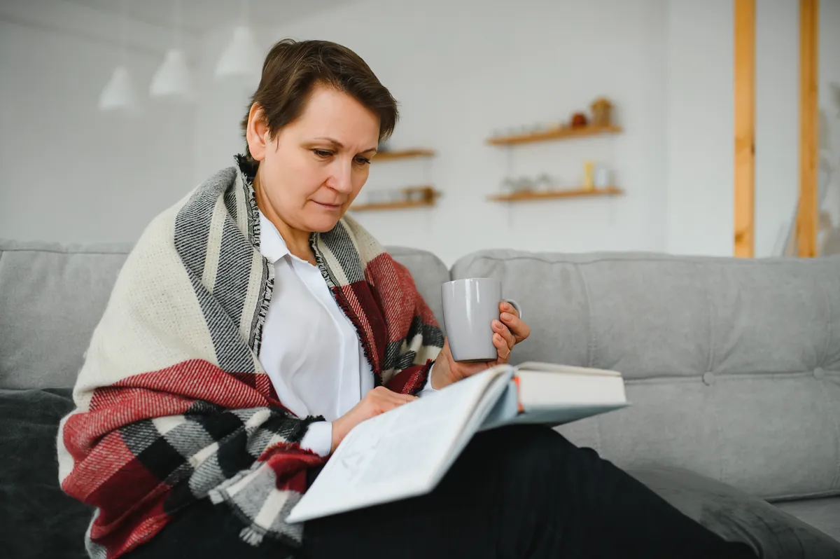 Woman wrapped in a red, white and grey plaid blanket on a grey couch, holding a mug and reading an open book