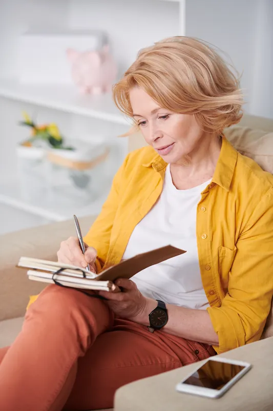 Woman with short strawberry-blonde hair and a yellow shirt writing in a notebook on a sofa, with her phone on the armrest