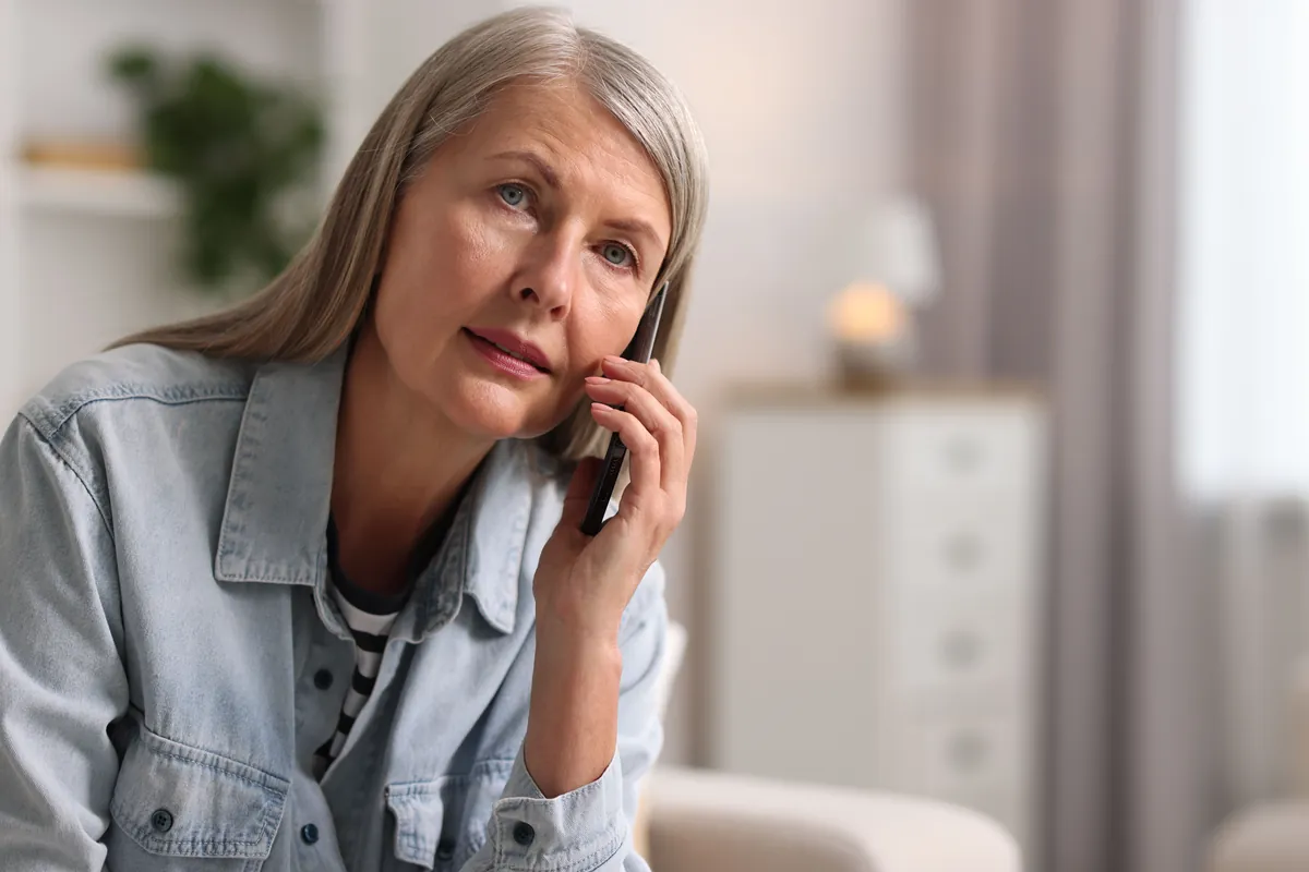 Woman with long grey hair and a light denim jacket talking on her phone at home with a worried expression